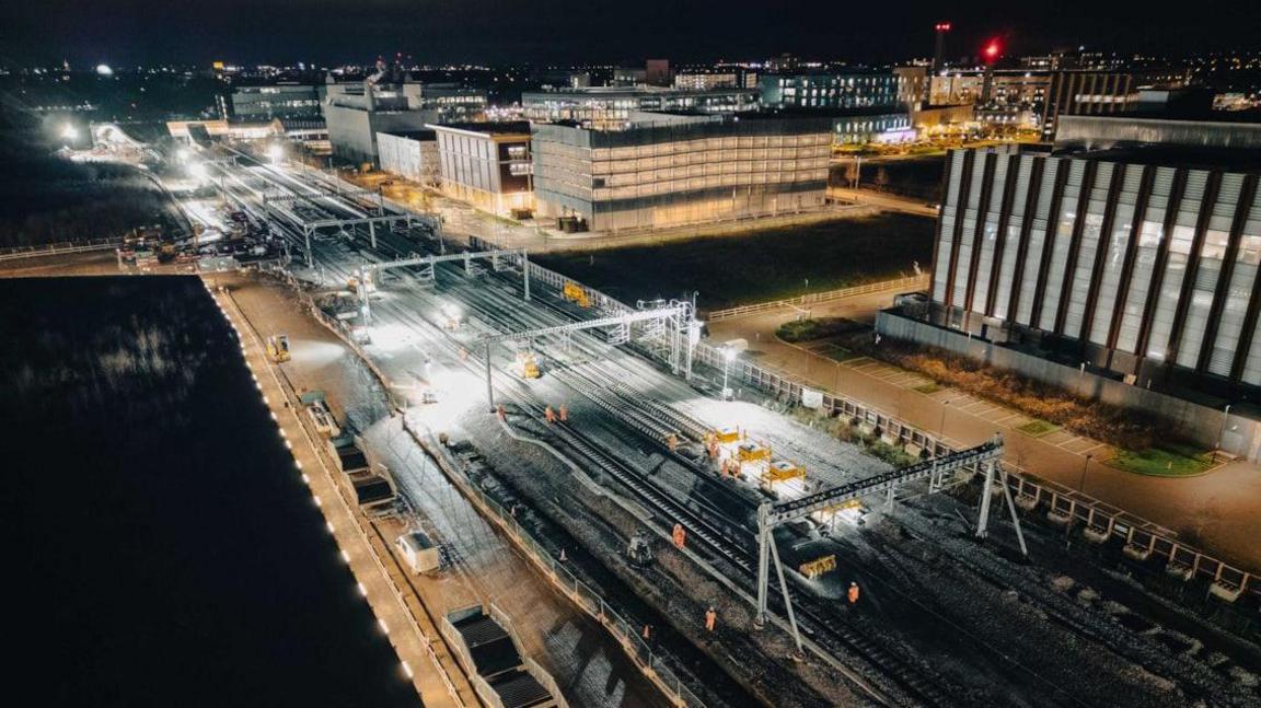 Aerial view of Cambridge South station at night-time. There are construction workers on the railway tracks.