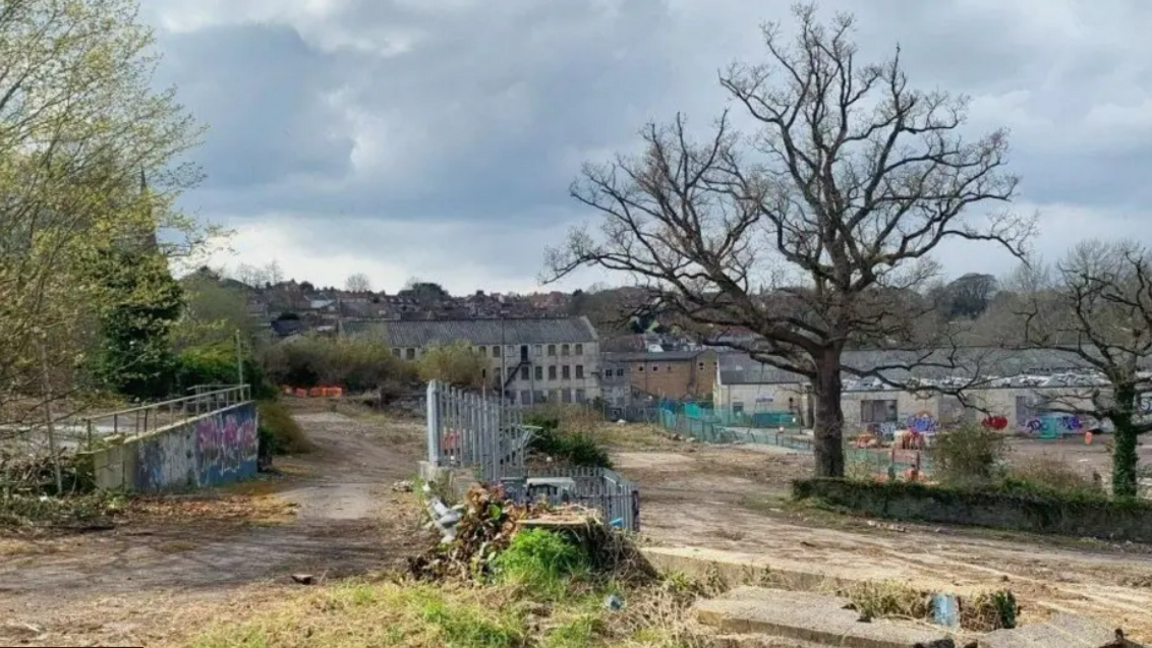 Abandoned site in Frome looking derelict, it has old fences, scrubs and houses in the background. 