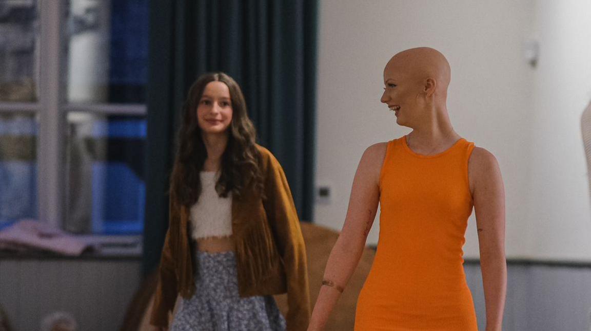 Enlli Thompson taking part in  a fashion show for the charity, Tenovus Cancer Care. Her head is shaven and she is wearing a short sleeve orange dress. To her right is another young girl who is also taking part in the fashion show. She is wearing a brown jacket, white crop top and a purple floral skirt.