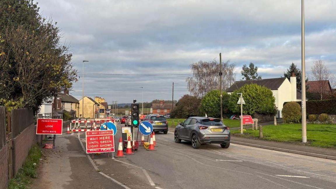 Temporary traffic lights, road signs and orange cones on the A49. There are cars driving through a green light on the right hand side of the road. In the distance, the yellow painted pub is visible.