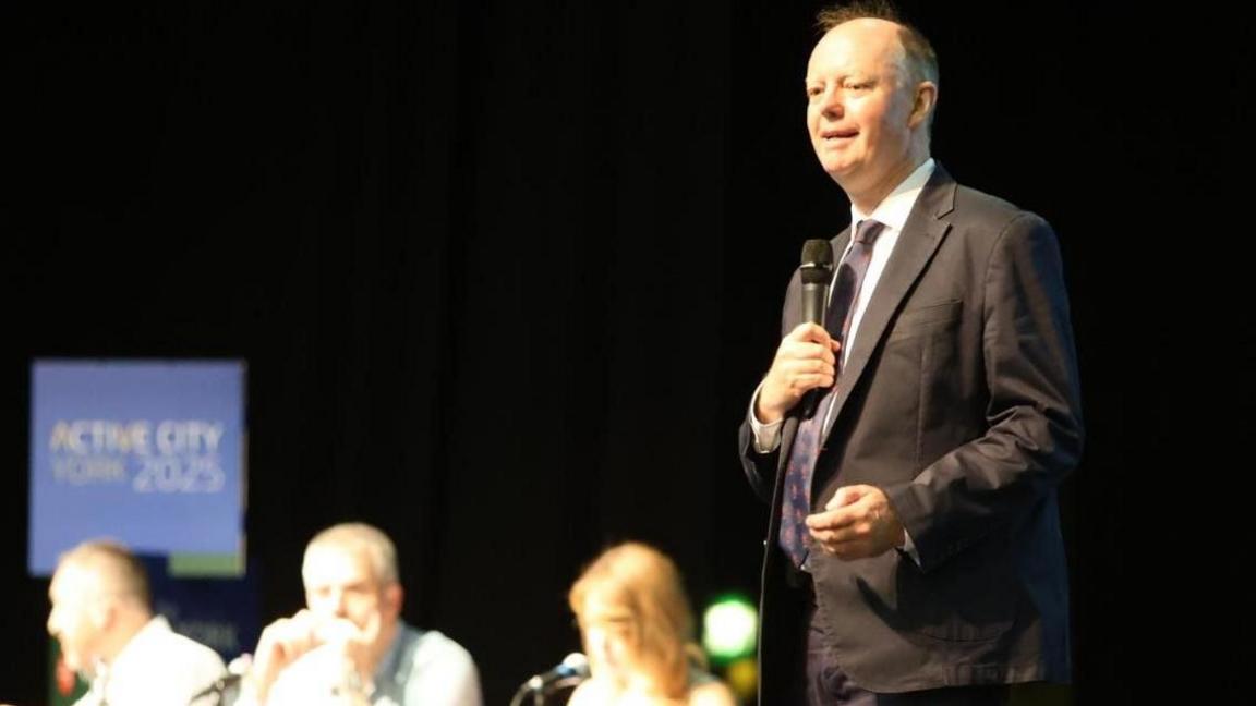 A man in a dark suit wearing a tie and holding a microphone stood on a stage in front of a table of delegates.
