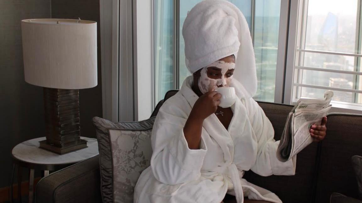 A woman is sat on a sofa in a white bath robe, having a cup of tea whilst wearing a face mask and reading the newspaper. 