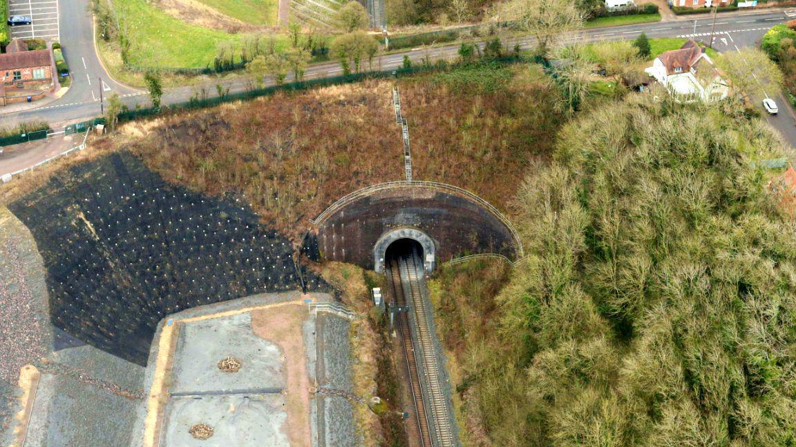 A view of the other side of the tunnel in March 2022. The aerial view shows the steep sides of the cutting and buildings dotted around the tunnel. A road goes over the tunnel and there are junctions on both sides.
