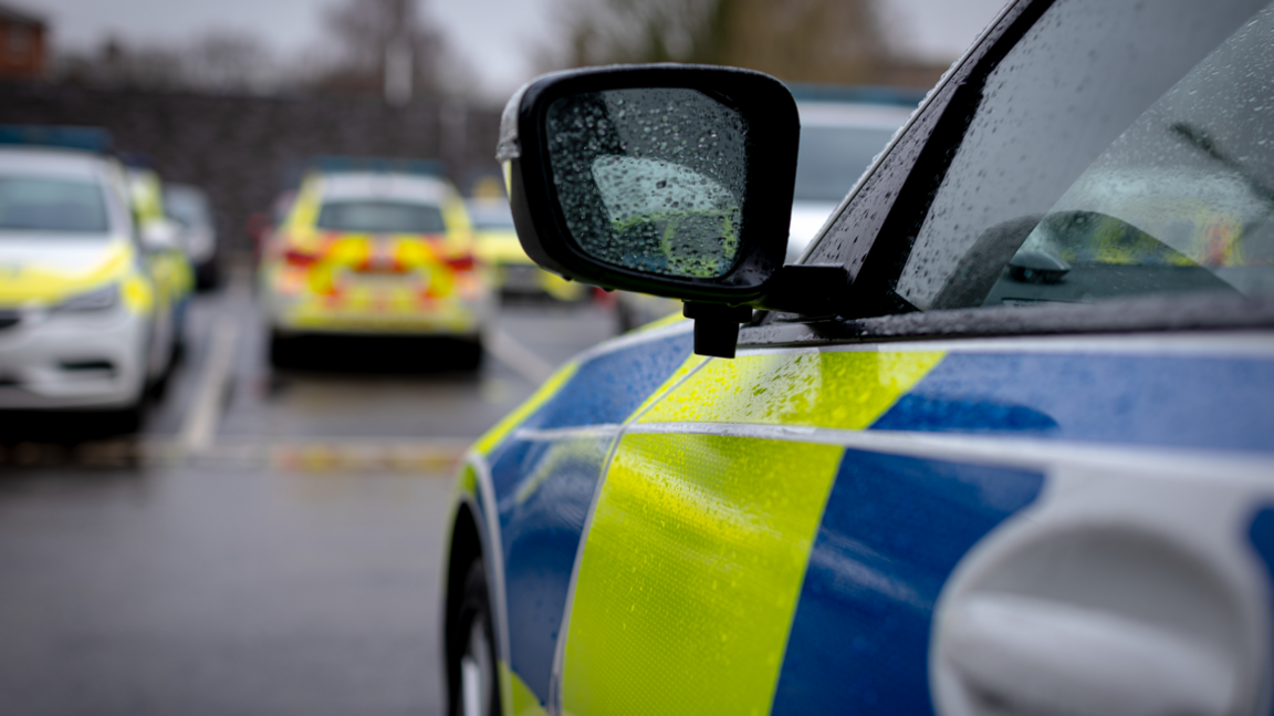 A photo taken from the passenger side handle of a police car, facing forwards. The car is wet from the rain. The wing mirror is the main focal point and it has water droplets on the glass. The car has alternating yellow and blue squares. In the distance are blurred police cars parked in parking spaces. 