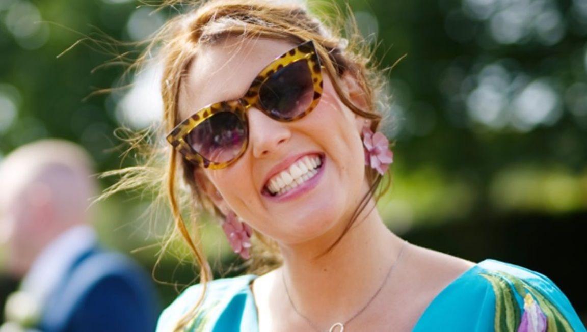 Mary has a beaming smile. Her light brown hair is blowing in the wind and she is wearing sunglasses, a colourful summer dress and pink flower earrings.