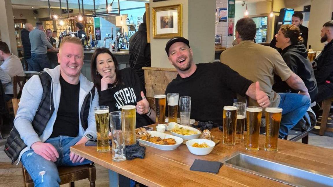 Three people sat at a pub table, smiling and with thumbs up, with numerous beers in front of them