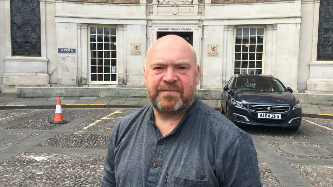 Bill Revans, in a grey casual shirt standing outside county hall in Taunton with a large black car just behind him