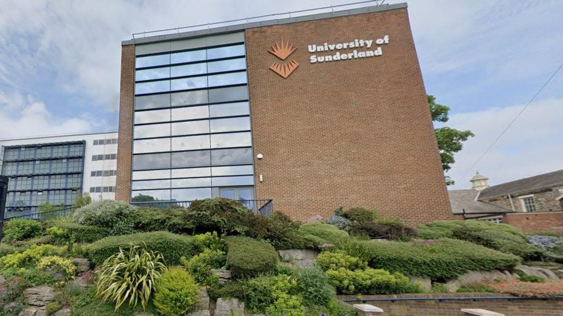 Part of Sunderland University's campus in the city centre. It shows a brown brick building, with the orange logo of the university. There are some landscaped shrubs in front of it. There is another building. mostly made of glass, further back to the left. 