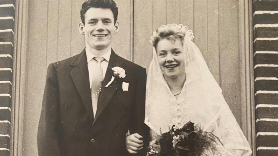 A black and white image of a bride and groom, Brian and Isobel Teasdale, in front of a church. She is wearing a veil and is holding a bunch of flowers and foliage. He has on a suit with a carnation in his buttonhole. Both are smiling.