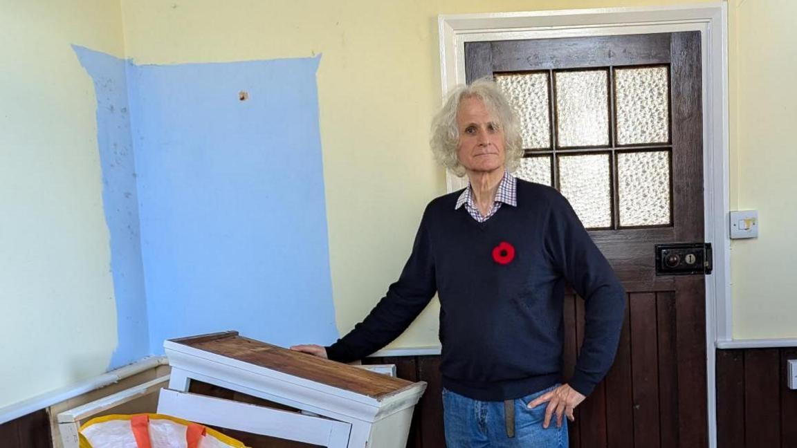 Eric Fenwick, who has long, grey hair and is wearing a navy jumper with a poppy and jeans, standing next to a broken wooden cupboard.