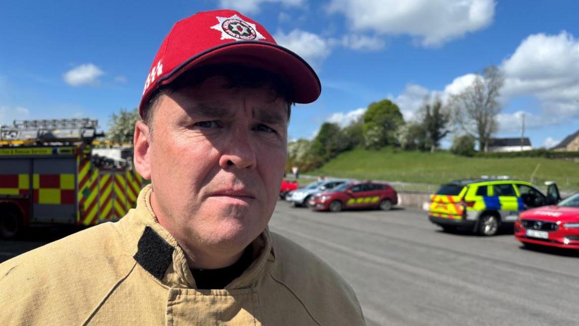 David Doherty has short dark hair and is wearing a red cap with the Northern Ireland Fire and Rescue badge on it. He is wearing a brown firefighter suit. Behind him is a yellow and red fire engine and a yellow and blue police car along with other vehicles.