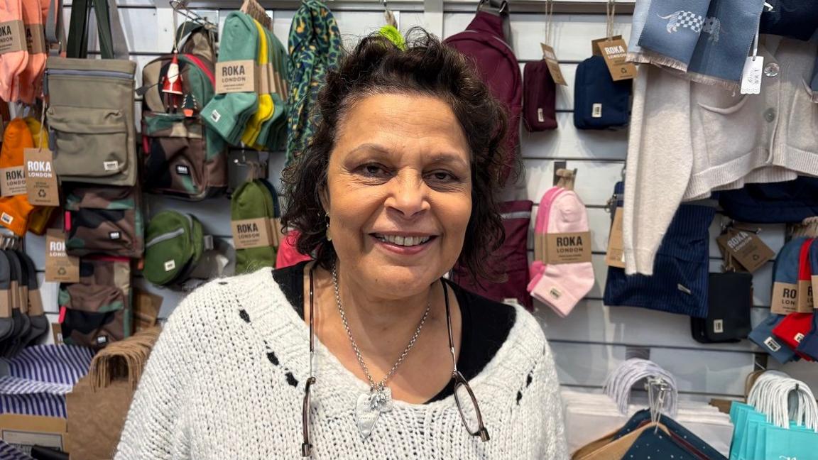 Caroline Gates with long black hair wearing a white sweater and split glasses on a string. There are clothes and bags on display behind her.