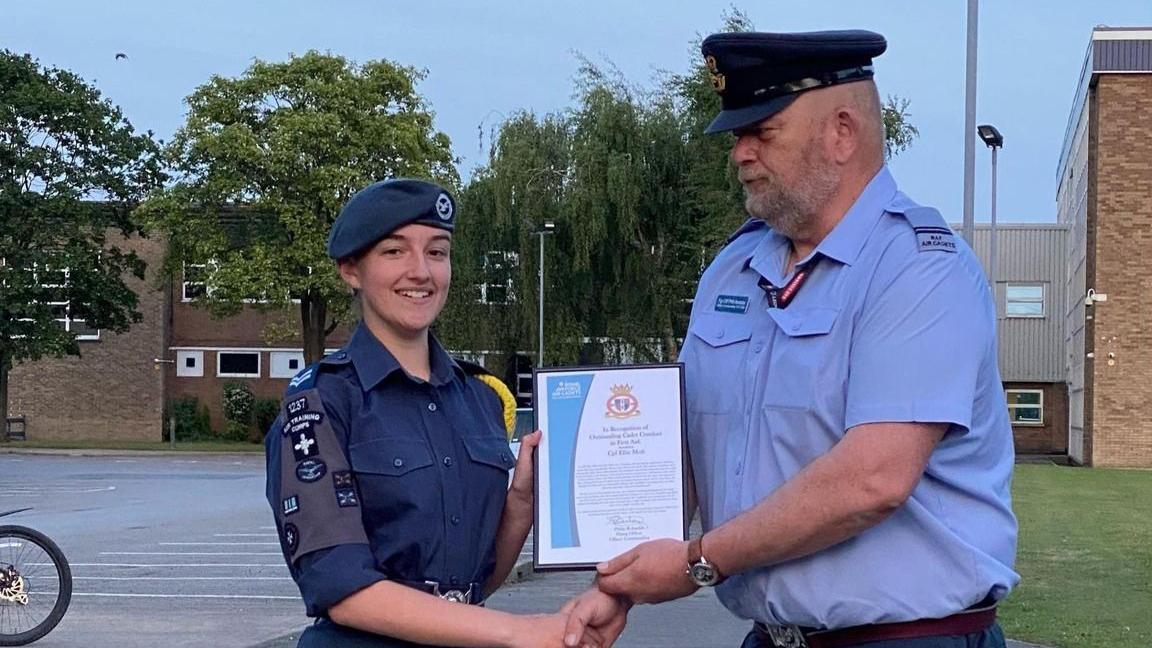 A teenage girl dressed in a navy RAF cadet uniform and a beret is smiling at the camera whilst holding a framed certificate and shaking the hands of a man dressed in a RAF uniform. They are stood in a car park in front of buildings.