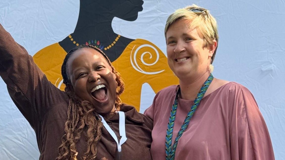 Two people smiling and posing together in front of a colourful background featuring artwork of a silhouetted woman.