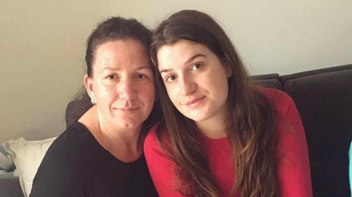 Una is sitting on a sofa next to her mother. Una is wearing a red top, while her mother is wearing a black top.