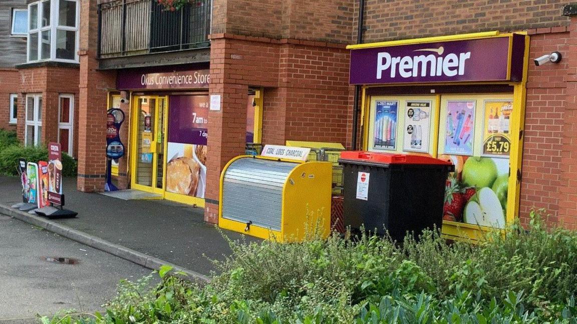 The front of Okus Convenience Store with its purple and yellow branding, with bins and pavement in front 