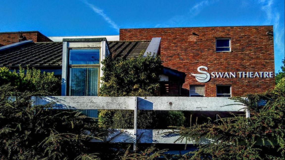 The outside of the Swan Theatre in Worcester. It is a brick building with shrubs planted around it. The theatre sign includes an image of a swan. It is a sunny day with a blue sky.