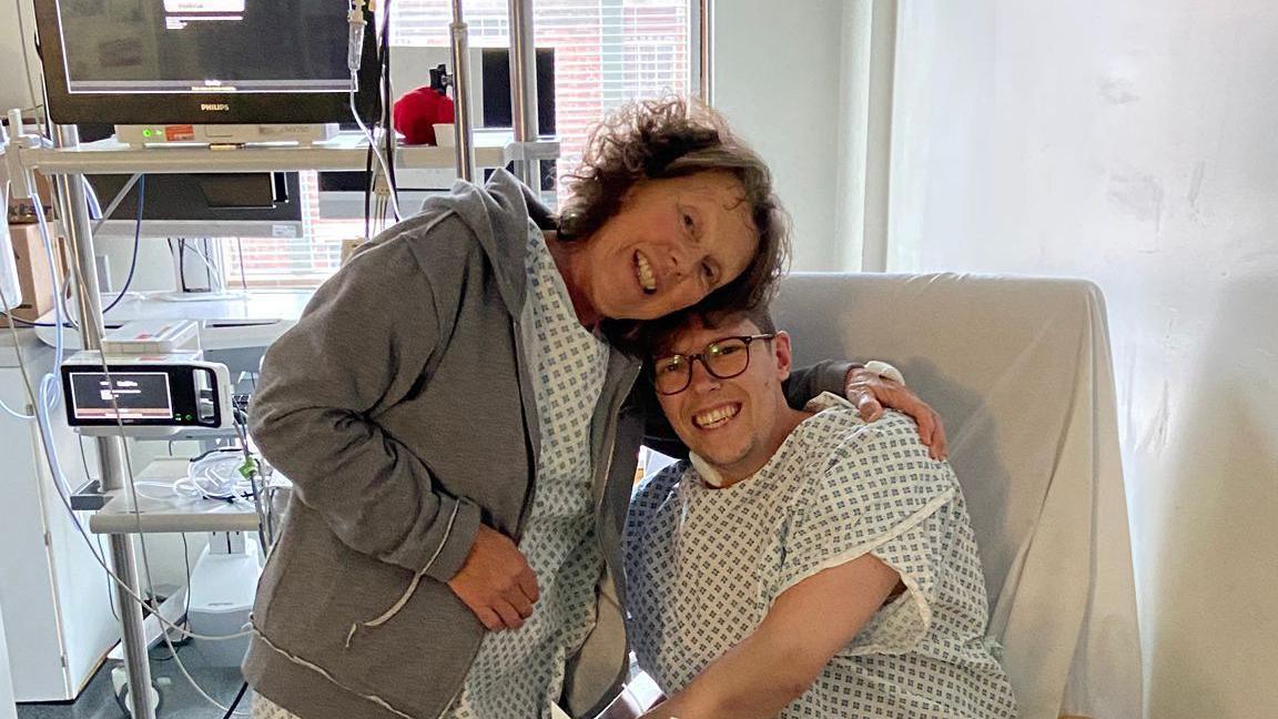 A woman leaning towards a younger man who is sitting in a hospital chair. The man is smiling and wearing a hospital gown. The woman is also wearing a hospital gown, which is partly covered by a grey zip hoodie. Medical equipment can be seen in the background. 