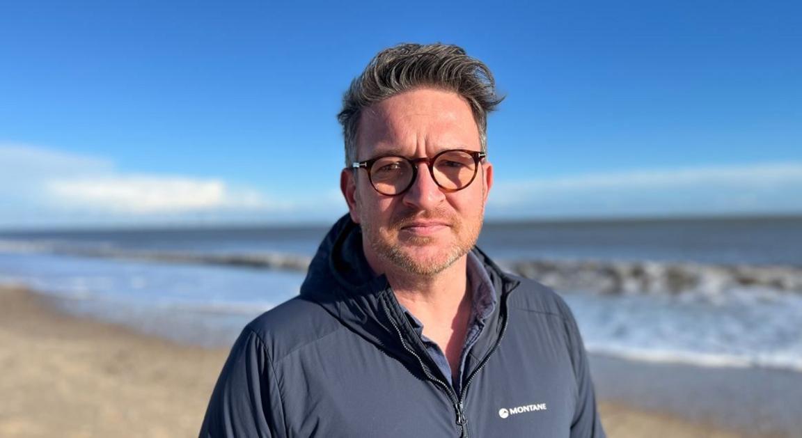 Man with glasses and beard standing beside the sea, looking at the camera