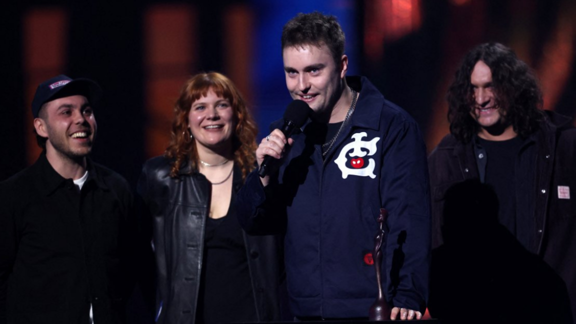 Sam Fender speaks as he receives the award for Best Rock/Alternative Act, at the Brit Awards. He wears a navy jacket with a white logo and three band members stand beside him.