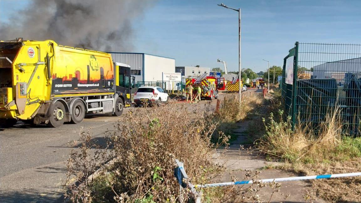 A blue and white police cordon on site of the fire- with a yellow refuse lorry and fire trucks in front of it