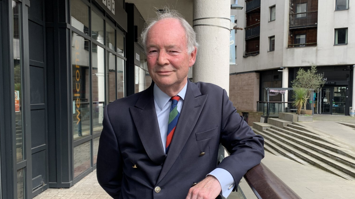 Philip Seccombe leans on a bannister outside of a BBC studio. He has white hair on the sides of his head and is bald on top. He is wearing a black suit jacket, a multi-coloured tie and a light blue shirt.