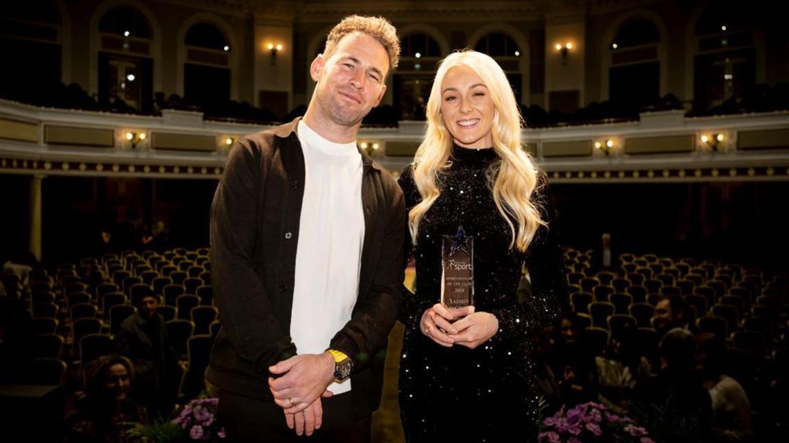 Sir Mark Cavendish and Yasmin Ingham standing on the Villa Marina stage smiling. Ingham has long blonde hair and is wearing a sparkly long black dress and is holding her award. The auditorium behind them is a sea of empty chairs and is dimly lit.