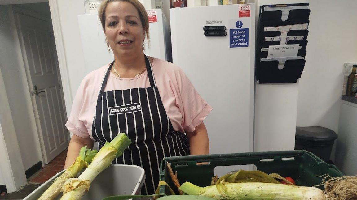 Raja El Faleh who has tied-back dark hair, is wearing a black and white apron and is standing behind a box of vegetables.