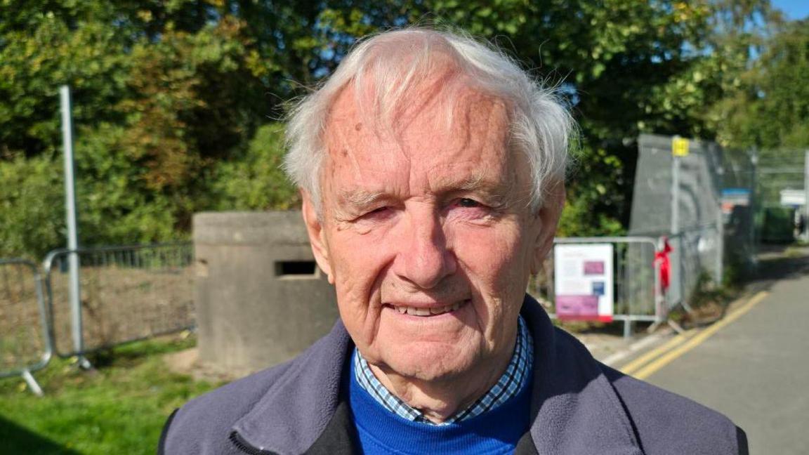 Bill Morgan is an elderly man with white hair. He wears a fleece jacket, checked shirt and blue jumper. He is smiling, and stands in front of the pillbox.
