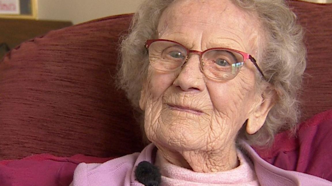 Dorothy Howard is sitting in a chair and wearing a pink fleece and t-shirt. She has medium length white hair and pink-trimmed glasses.