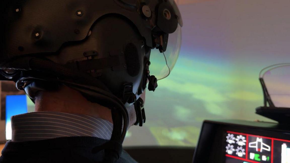 A man wearing a black helmet with a clear visor. He sits in front of a flight simulator screen with controls in front of him.