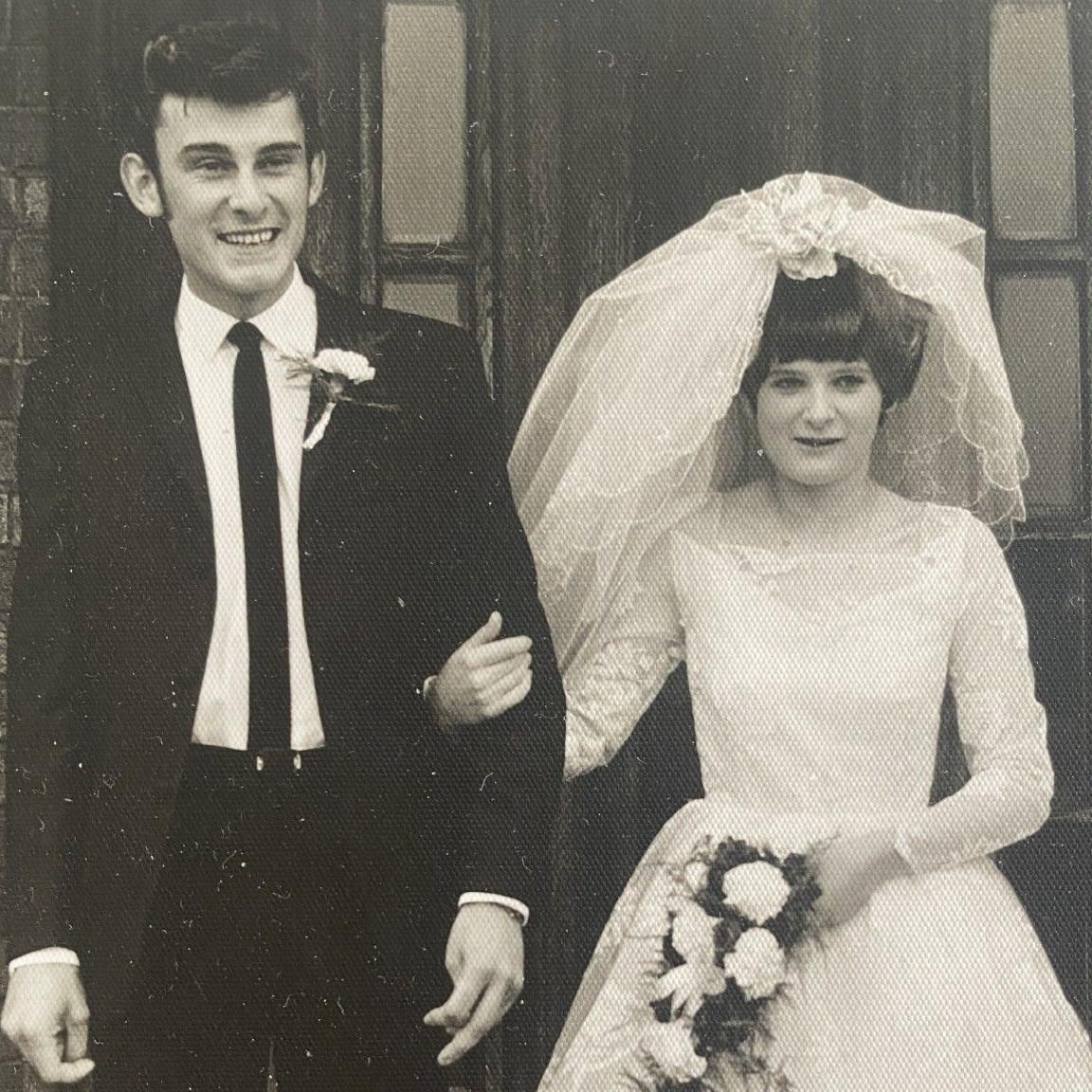 A black and white photo of Ron and Eileen on their wedding day. Ron is on the left wearing a suit while Eileen, on the right, is wearing a white dress with a veil swept over the back of her head. She is holding a bouquet of flowers in one hand and holding Ron's arm with the other