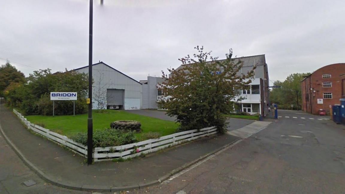 Two metal corrugated buildings stand on a corner street, there is a tree in front and to the left. A sign that reads 'Bridon' is positioned in grass on the left.