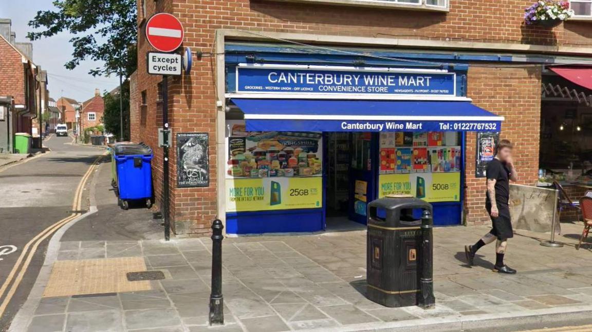 A Google image of Canterbury Wine Mart. It is a red brick end-terrace building with a blue awning. 