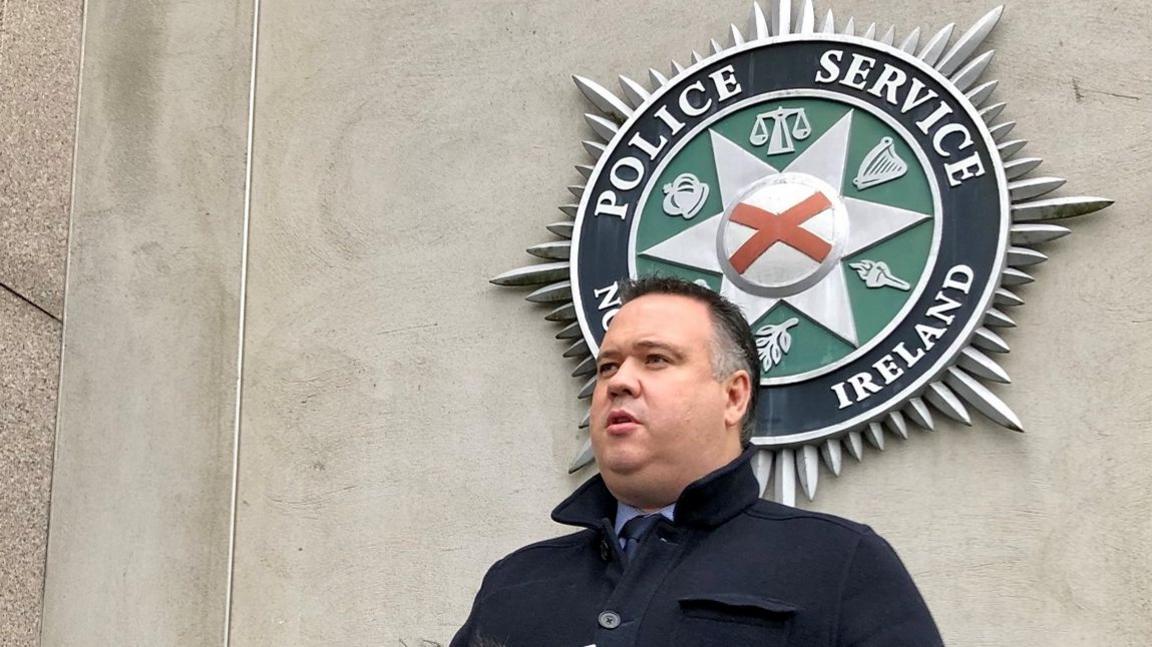 A man with dark hair in front of a Police Service of Northern Ireland logo