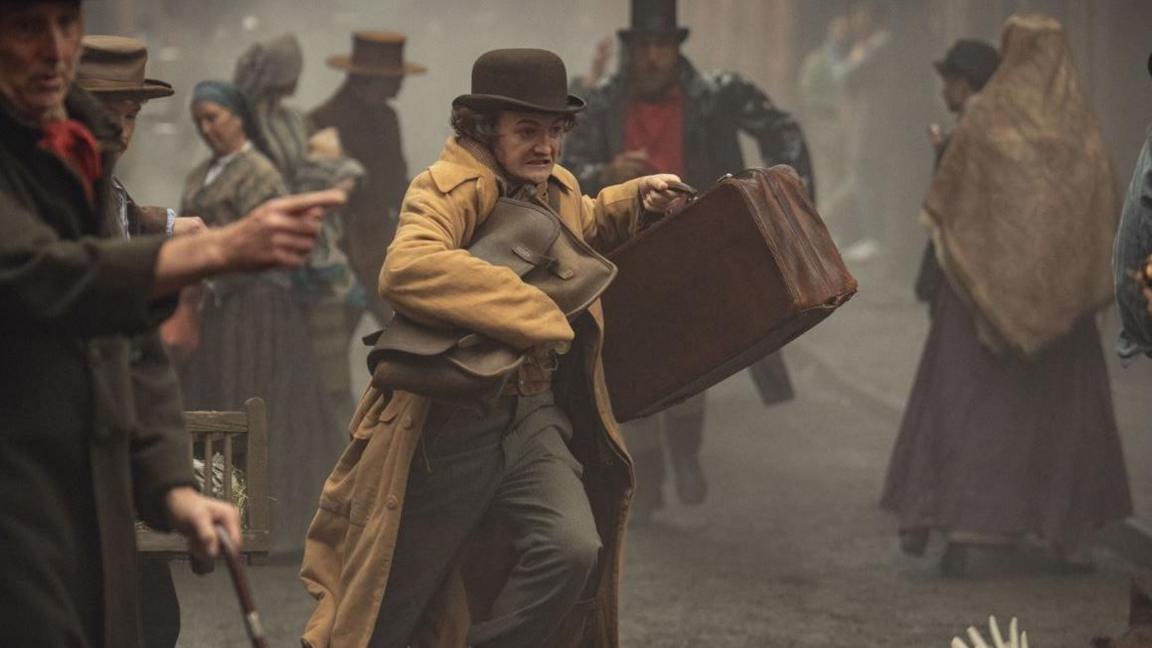 Jack Gleeson as Byron Hedges wearing a yellow coat and brown cap holding two bags and running in the street