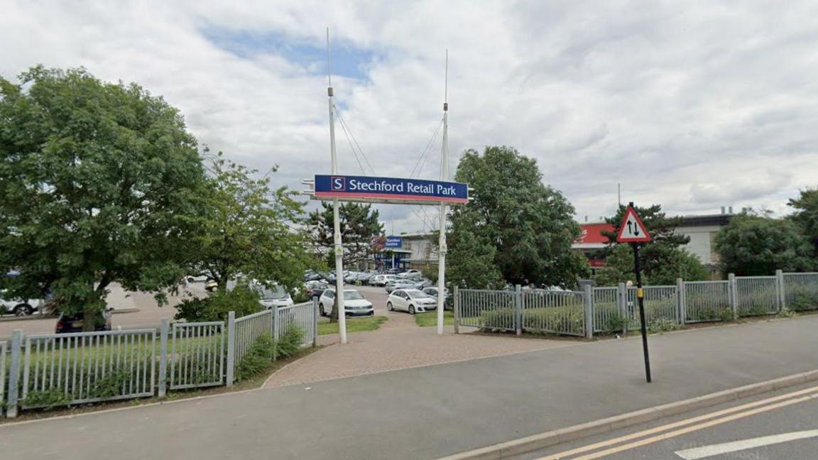 A Google street view image showing the entrance to a retail park on a pavement next to a main road. A blue, red and white sign above the entrance says "Stechford Retail Park". There are grey fences and trees either side. Beyond the entrance there is a car park and a line of large shops.