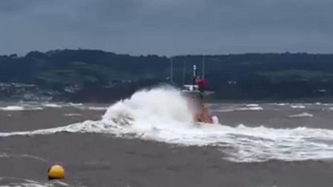 A partially occluded lifeboat at sea with a yellow buoy in the foreground and a large wave between it and the lifeboat in a greyish sea with land in the background of a rural and partially wooded nature above which is a leaden sky filled with foreboding and mystery.