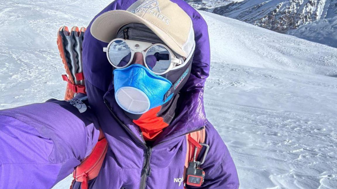 Adriana Brownlee stands and looks towards the camera in a purple snowsuit and brown hat, while wearing a blue mask and polarised glasses, with snow behind her