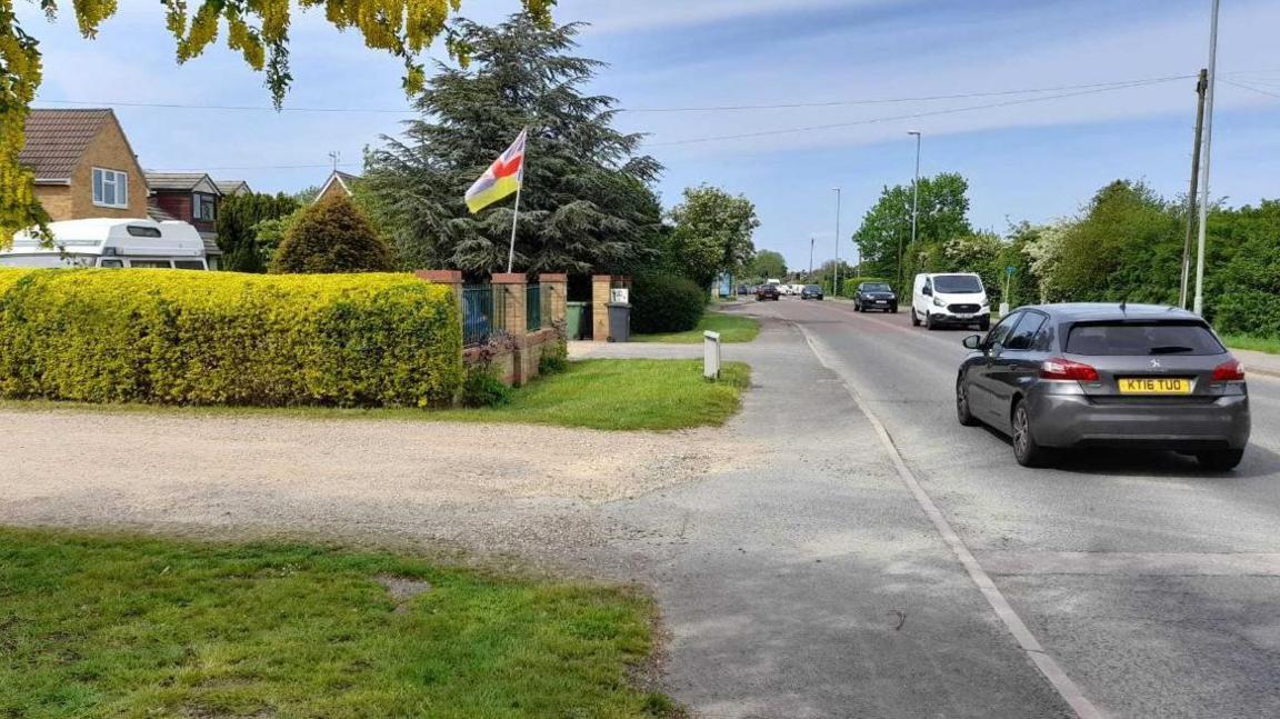 An entry to a gravel road, with grass on either side of the gravel. On the right is a single-carriageway road with cars and vans travelling in both directions. On the far side of the gravel road is a hedge with house-roofs and a campervan roof above