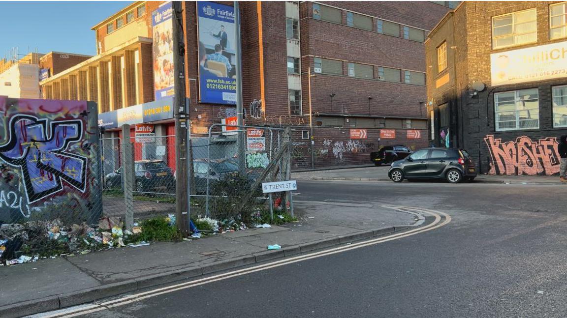 The corner of two roads. A street sign reads "Trent Street". A wire fence runs along the edge of the pavement, and a nearby brick wall is covered in graffiti. A building on the other side of the road has also been graffitied.