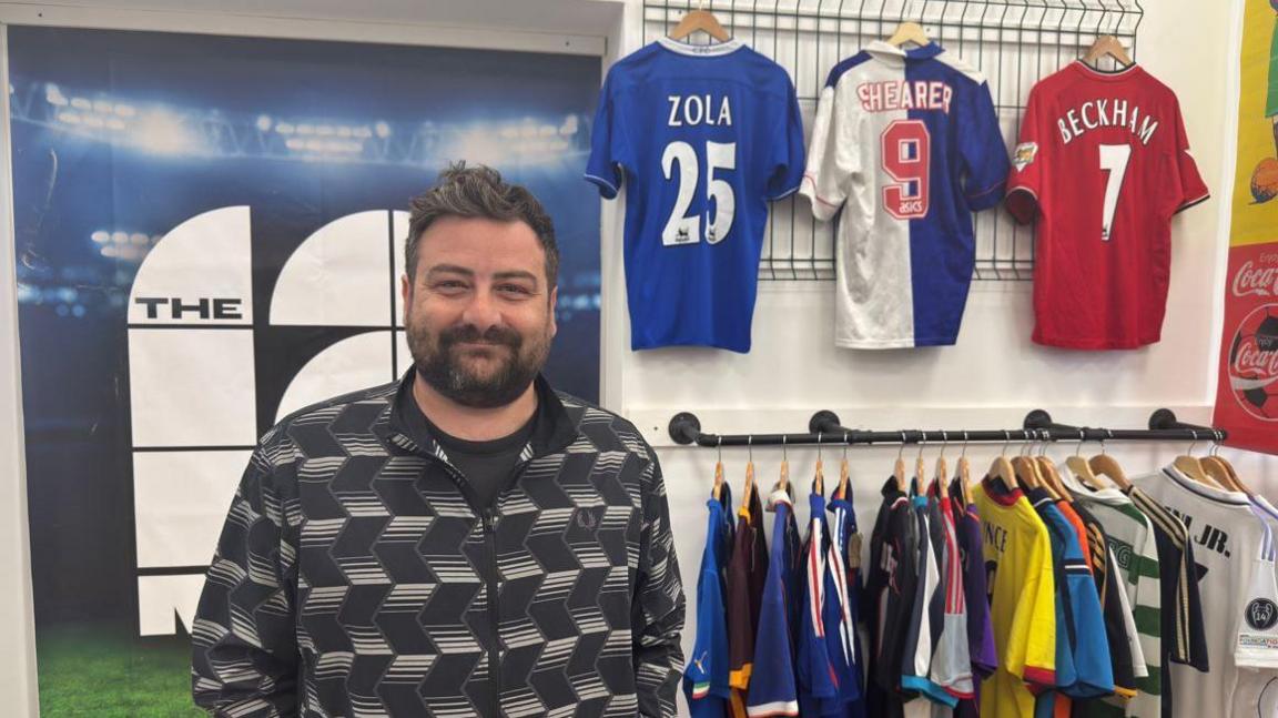 A man with dark hair and a beard is wearing a black shirt with geometric white patterns. He is standing in front of two racks of brightly coloured football shirts with three on display in the upper rack.