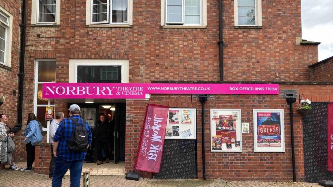 Two storeys are visible in this photo taken from the outside of the theatre. A pink and white sign above an entrance says Norbury Theatre & Cinema. Five people are standing outside.
