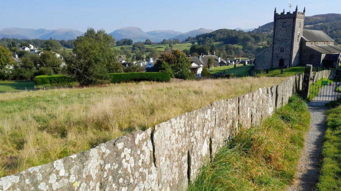 Tall shards of slate, which are covered in lichen and moss, interlock together together on farm land, marking a boundary. There is a front part of a digger tied to one shard, after it was just laid into the ground. 