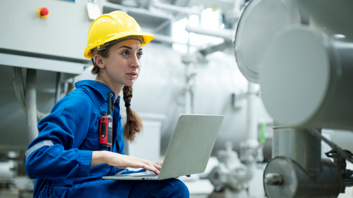 Young female engineer wearing yellow hard hat and blue overalls, working on a laptop at industrial site 