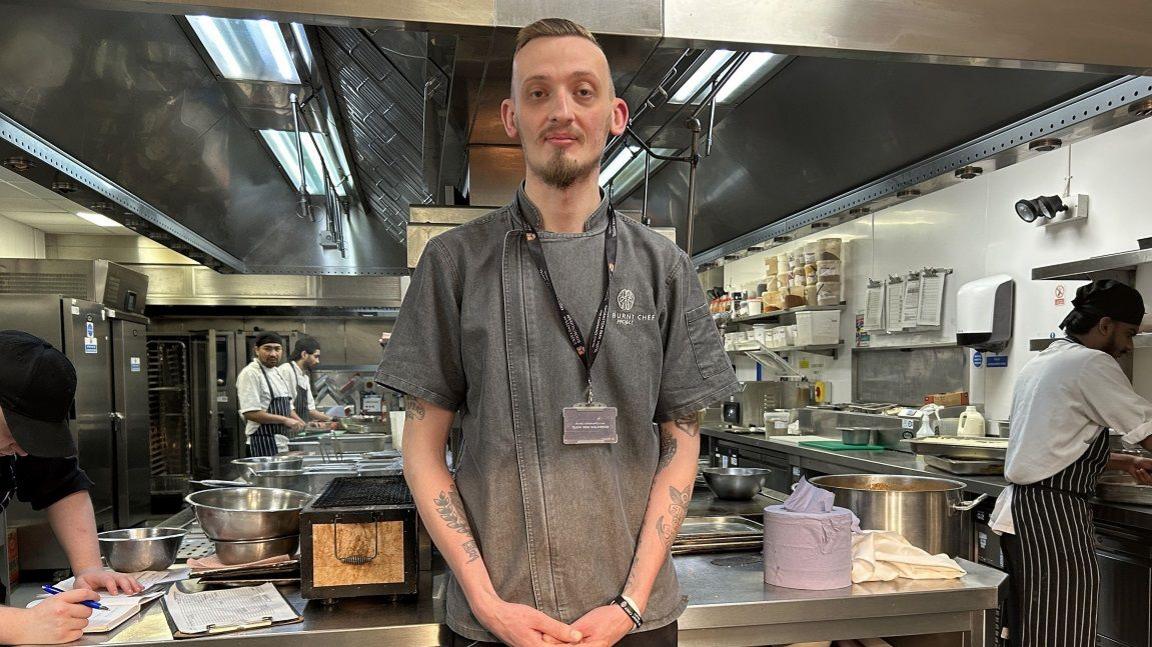 A man stands in a kitchen. He has short brown hair and facial hair. He is wearing a grey denim shirt with a logo on it. In the background are several chefs with stripey black and white aprons on