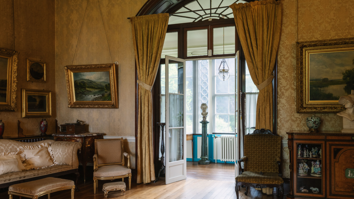 One of the rooms in Preston Manor on the outskirts of Brighton. This room has a number of gold-framed photographs, chairs and a sofa. White double doors can be opened out into the corridor. 