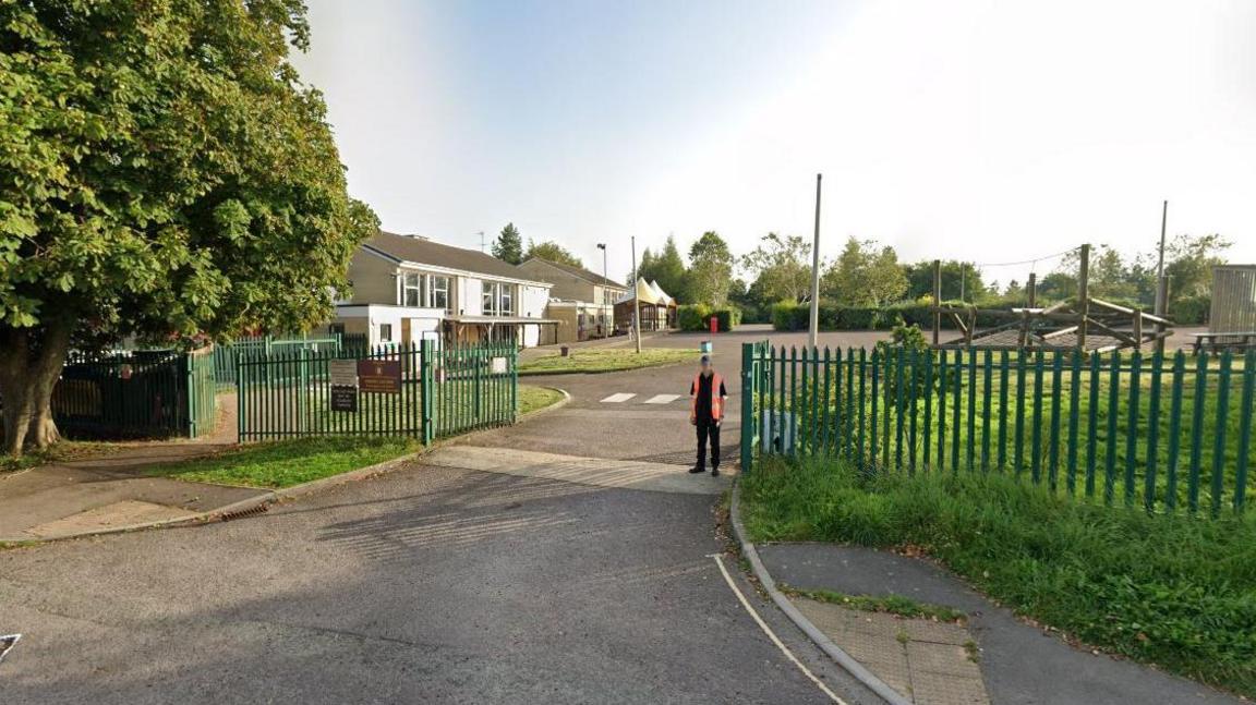 Nailsworth Church of England Primary entrance on a sunny day - one storey buildings with a tarmac and grass playground, a large tree to the left of the gates.