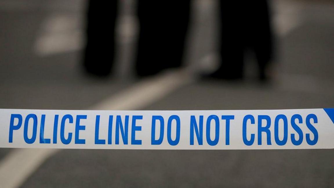 A close up of police tape that reads "police line do not cross". The blurred legs of two people are in the background.