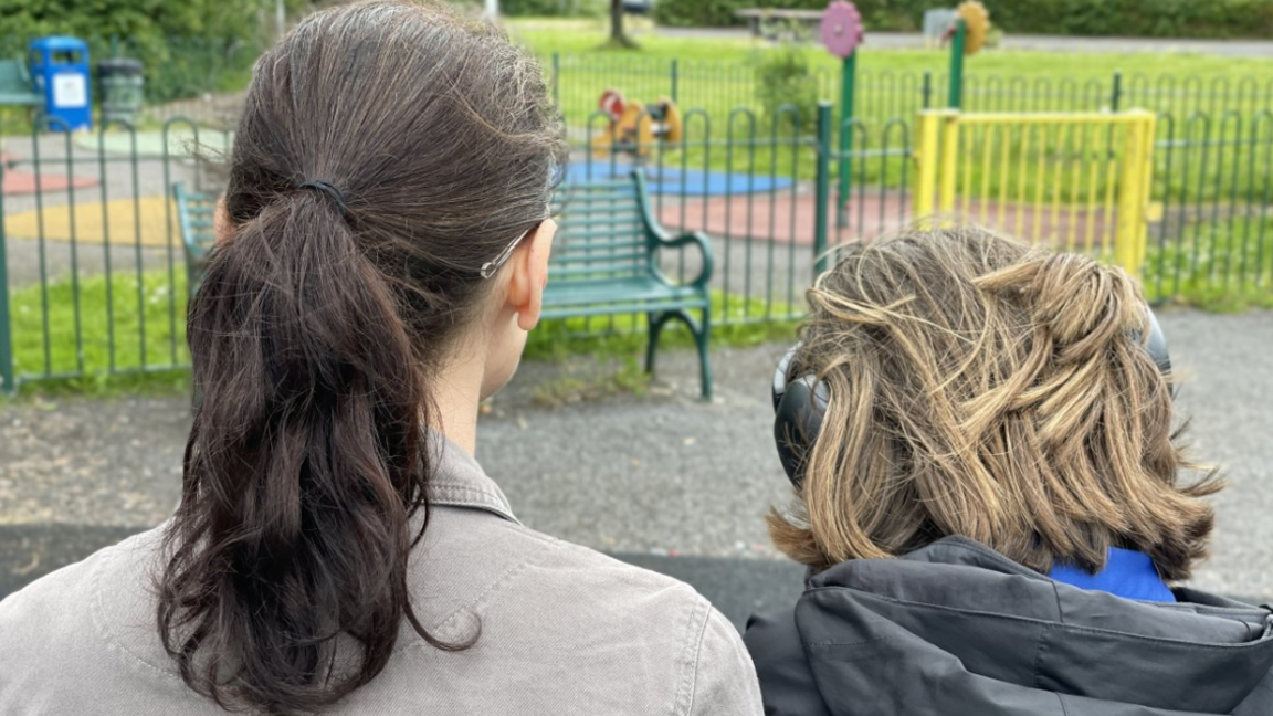 A child wearing headphones next to an adult, with just the back of their heads visible, sitting in a children's playground in a park